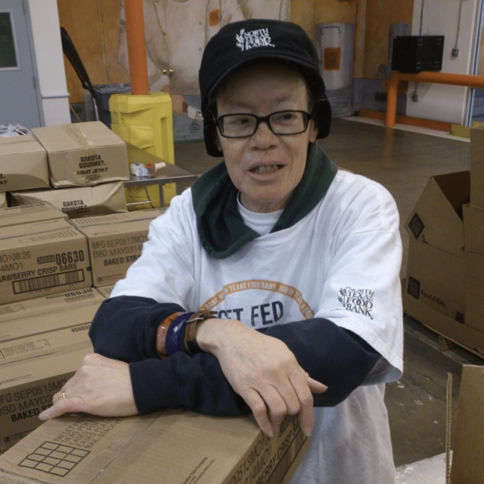 Mary Hinton Older woman standing amongst boxes with an North Texas Food Bank hat
