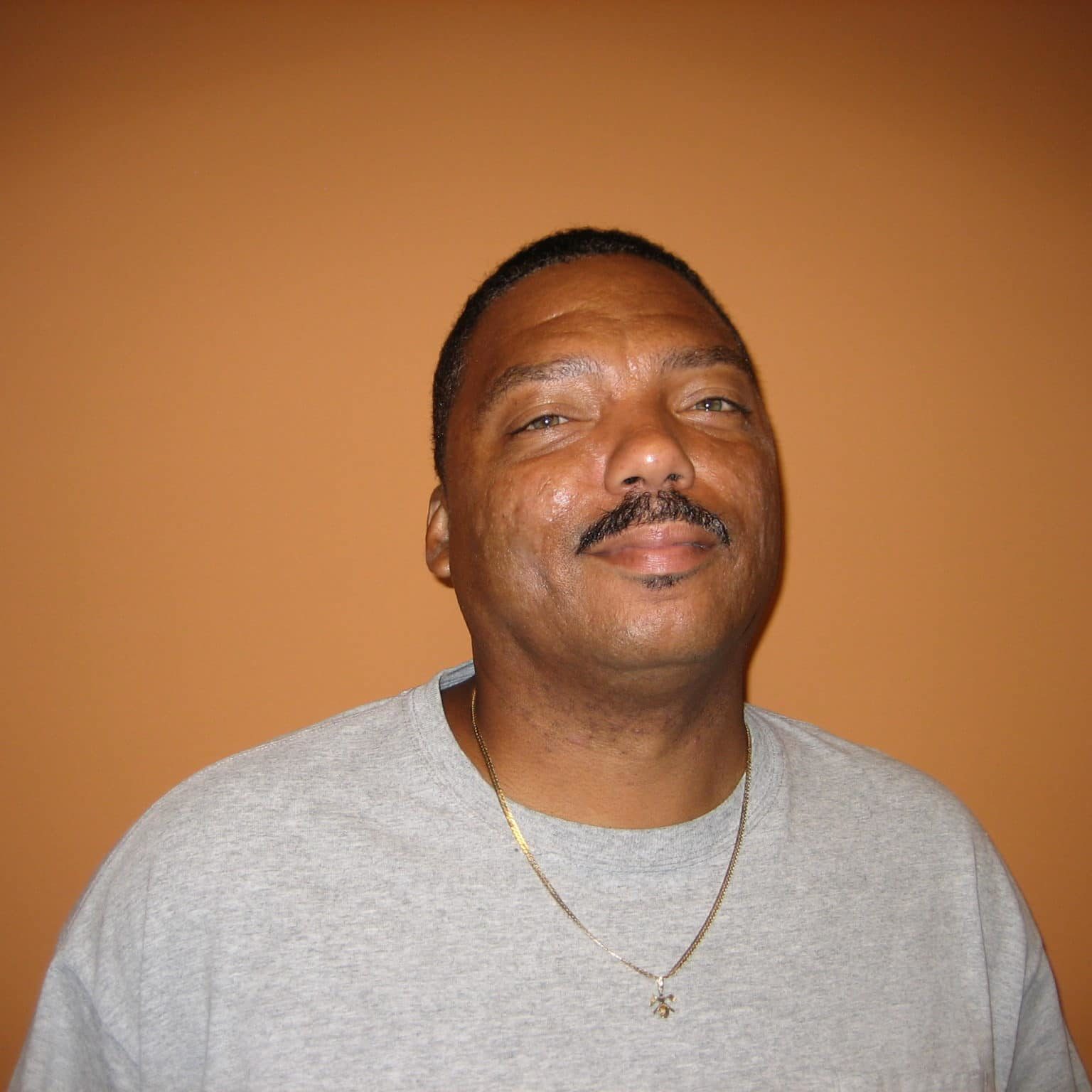 Vince Rhinehart Logistics Coord Vince Rhinehart in a grey t-shirt and gold necklace in front of an orange background